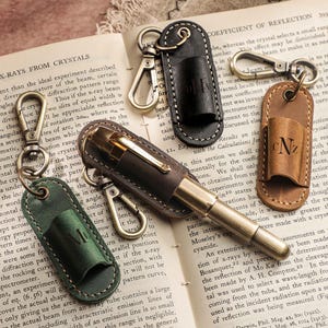 Könnte beinhalten: Eine Sammlung von Leder-Schlüsselanhängern und einem Stifthalter, jeweils mit Metallverschluss. Die Schlüsselanhänger sind in Grün, Schwarz und Braun gehalten und mit Initialen versehen. Die Artikel liegen auf einem offenen Buch.