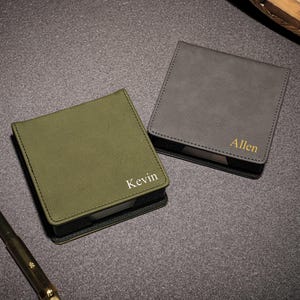 Può includere: Due blocchi per appunti quadrati personalizzati. Uno è verde oliva con "Kevin" in testo bianco, l'altro è grigio con "Allen" in testo dorato. Una penna oro e nera poggia sulla superficie. I blocchi per appunti sono su una superficie grigio scuro.