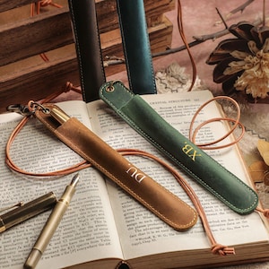 Puede incluir: Estuches de cuero para bolígrafos en marrón y verde, con un bolígrafo dorado y un libro en el fondo. Los estuches tienen un cordón de cuero y algunos tienen iniciales. El libro está abierto en una página con texto.