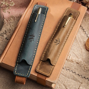 Op de afbeelding: Twee leren penhouders, één in teal en één in bruin, elk met een gouden pen. De teal houder heeft de initialen "ML" in reliëf, de bruine een takontwerp. Beide zijn gestikt met crèmekleurig garen en hebben een bruine elastische lus.