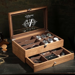 May include: A wooden watch and accessory box with two drawers. The top drawer holds several watches, while the bottom drawer contains sunglasses and other items. The box has a glass top with a decorative design.