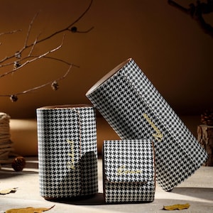 May include: Three cylindrical storage cases featuring a black and white houndstooth pattern. Each case has a brown interior lining and gold cursive text embroidered on the exterior. The set includes one tall case, one medium case, and one small case.