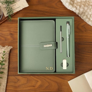 Könnte beinhalten: Ein salbeigrünes Schreibwaren-Geschenkset in einer passenden Box, bestehend aus einem strukturierten Notizbuch mit goldfarbenem Verschluss und den goldgeprägten Initialen N.D., einem passenden Kugelschreiber, einer Mine und einem kleinen rechteckigen Metallteil.