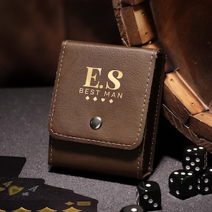 May include: A brown leatherette dice and card holder with the initials "E.S." and the words "BEST MAN" printed in gold. The holder is closed with a snap. Black dice and playing cards are visible.