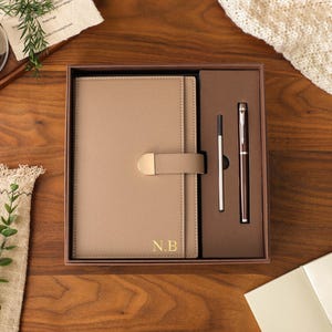 Op de afbeelding: Een beige notitieboek met ledertextuur, voorzien van een goudkleurige metalen sluiting en de goudkleurige initialen N.B. op de kaft, gepresenteerd in een bruine geschenkdoos met een bijpassende donkerbruine pen en zilverkleurige vulling.