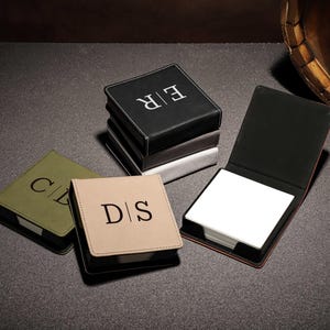 May include: A collection of personalized square notepads in various colors, including black, olive green, and tan. Each notepad features initials in a contrasting color. One notepad is open, revealing a stack of white paper.