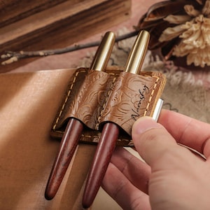 Puede incluir: Un estuche de cuero marrón para bolígrafos con dos bolígrafos. El estuche tiene un diseño floral en relieve y el nombre "Nashky". Los bolígrafos tienen puntas doradas y barriles de madera marrón oscuro.
