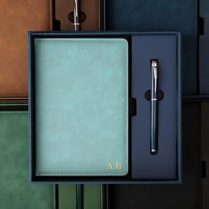 Op de afbeelding: Een lichtblauw rechthoekig notitieboek met de goudkleurige initialen A.B. in de rechterbenedenhoek, gecombineerd met een bijpassende donkerblauwe balpen. De set wordt gepresenteerd in een donkerblauwe geschenkdoos met een vak voor de pen.