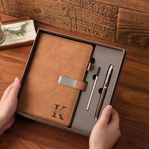 Könnte beinhalten: Ein beigefarbenes Notizbuch in Lederoptik mit silbernem Metallverschluss und dem eingravierten Buchstaben K sowie dem Namen KENT auf dem Einband, präsentiert in einer Geschenkbox mit silbernem Stift und schwarzer Mine in einer grauen Schaumstoffeinlage.