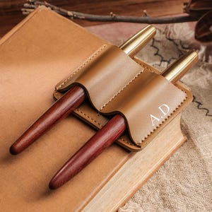 Puede incluir: Un portalápices de cuero marrón con dos bolígrafos. El soporte está grabado con las iniciales "A.D." y descansa sobre un libro cerrado. Los bolígrafos tienen un cuerpo de madera oscura y puntas de metal dorado.