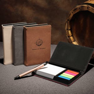 Könnte beinhalten: Ledergebundene Notizbücher in Beige, Grau und Braun, eines davon geöffnet mit Haftnotizen und einem Stift. Das braune Notizbuch hat ein Kompass- und Bergdesign mit dem Text "Journey of Adventure."