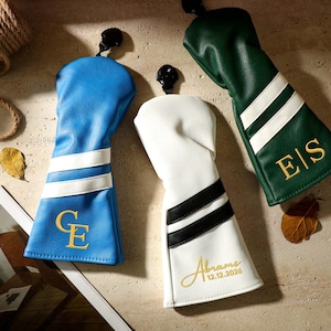 Könnte beinhalten: Drei Golfschlägerhauben aus blauem, weißem und grünem Kunstleder. Die blaue Haube hat weiße Streifen und goldene Initialen CE. Die weiße Haube hat schwarze Streifen und den goldenen Schriftzug Abrams 12.12.2026. Die grüne Haube hat weiße Streifen und goldene Initialen E|S.