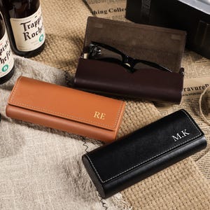 Könnte beinhalten: Eine Auswahl an Leder-Brillenetuis in Braun und Schwarz, mit personalisierten Initialen. Eine Brille liegt in einem offenen braunen Etui. Bierflaschen sind im Hintergrund, zusammen mit einer Schnurrolle.