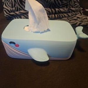 May include: A light blue whale-shaped tissue box with a white base and fins. The tissue box has a pink eye and a slot for dispensing tissues. White tissues are visible, emerging from the top of the box.