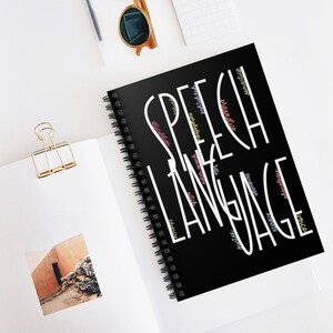 May include: A black spiral notebook with the word "SPEECH LANGUAGE" written in white. The words "articulation", "voice", "auditory", "grammar", "pitch", "language", "fluency", "morphology", "syntax", "prosody", "vocabulary", "speech", "rate", "disorder", "communication", "phonemic", "awareness", "phonology", "dialect", "sound", "rhythm", "intonation", "gestures", "nonverbal", "interaction", "social", "interaction", "meaning", "comprehension", "literacy", "reading", "writing", "expression", "ideas", "thoughts", "feelings", "knowledge", "culture", "society", "world" are written in various colors around the main text.