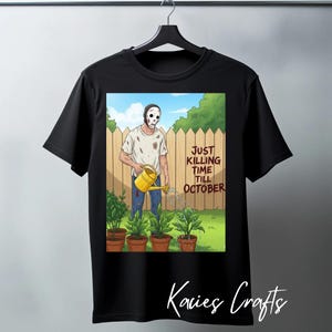 Könnte beinhalten: Schwarzes T-Shirt mit einer humorvollen Illustration einer maskierten Figur, die Pflanzen gießt. Der Text lautet "Just Killing Time Till October". Das Design befindet sich vor einem Hintergrund aus Holzzaun und grünem Laub.