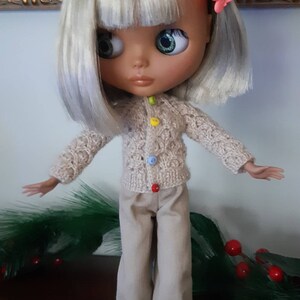 May include: A doll with blonde hair, wearing a beige cardigan with colorful buttons, beige pants, and white sneakers. The doll is standing on a white stand.