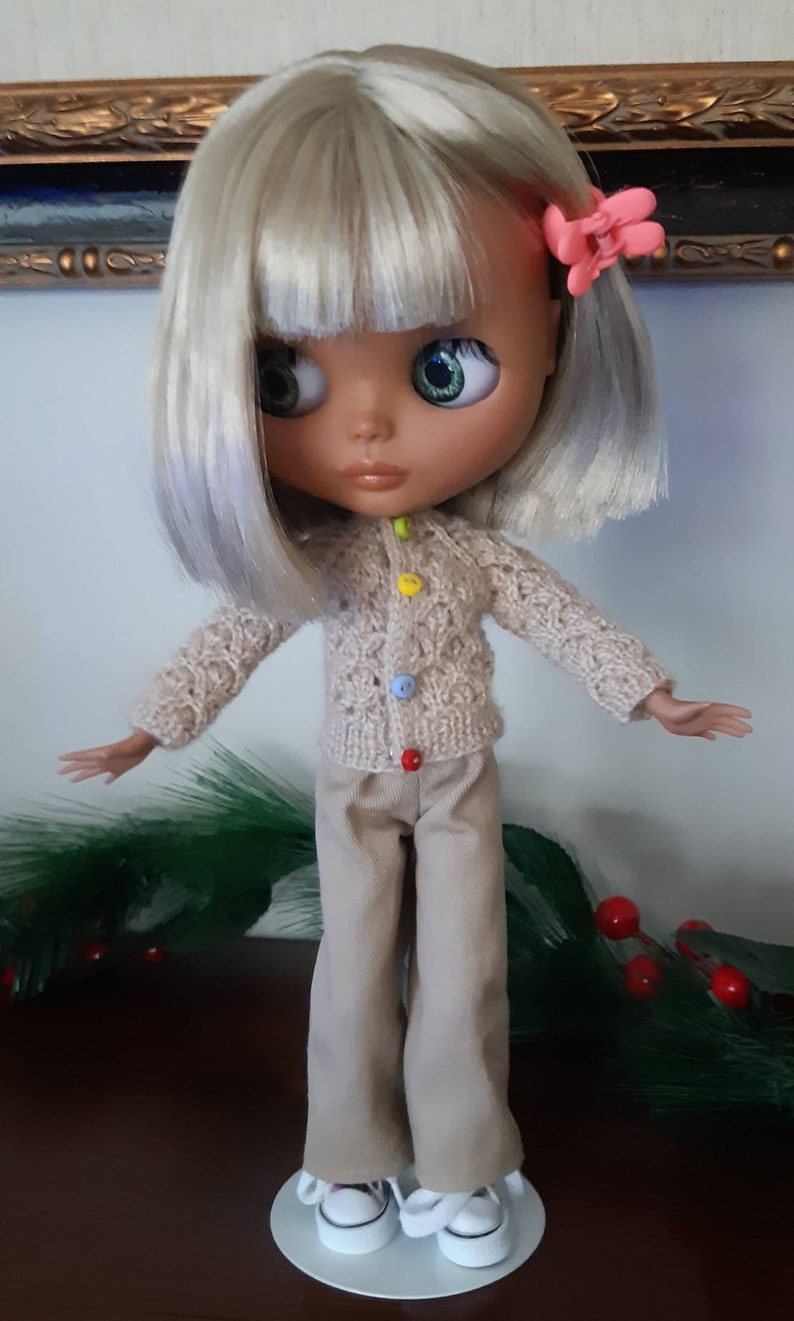 May include: A doll with blonde hair, wearing a beige cardigan with colorful buttons, beige pants, and white sneakers. The doll is standing on a white stand.