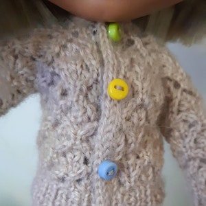 May include: A beige knitted cardigan with a button closure. The cardigan has four buttons in different colors: green, yellow, blue, and red.