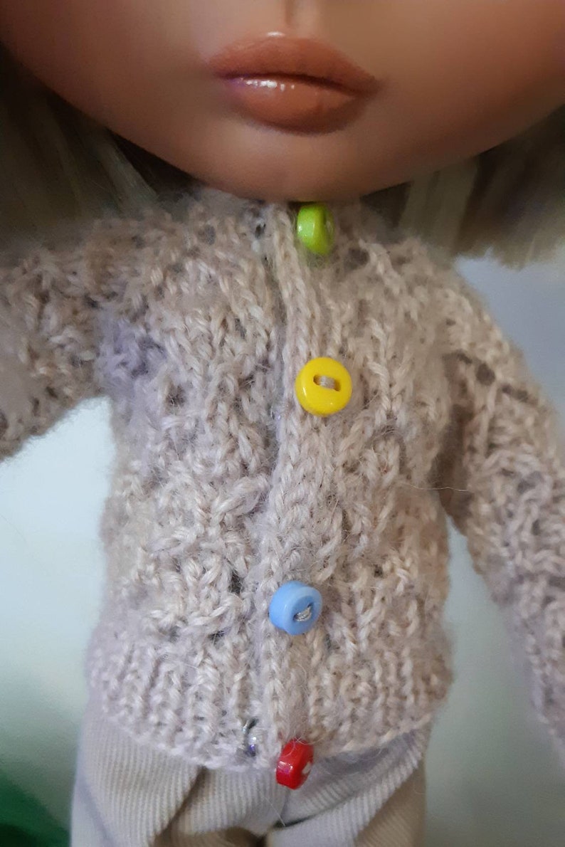 May include: A beige knitted cardigan with a button closure. The cardigan has four buttons in different colors: green, yellow, blue, and red.