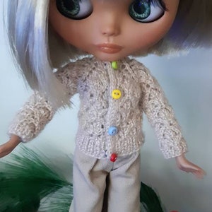 May include: A doll with blonde hair wearing a beige knitted cardigan with colorful buttons and beige pants. The doll is wearing white sneakers and has green eyes.