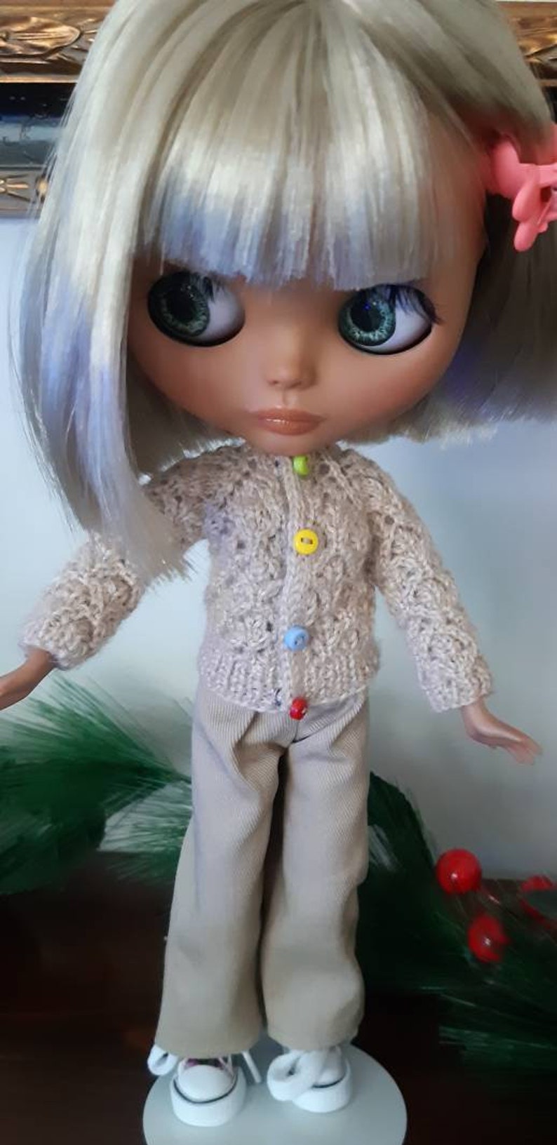 May include: A doll with blonde hair wearing a beige knitted cardigan with colorful buttons and beige pants. The doll is wearing white sneakers and has green eyes.