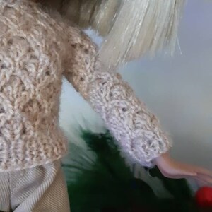 May include: A doll wearing a beige knitted sweater with a cable knit pattern and beige pants.