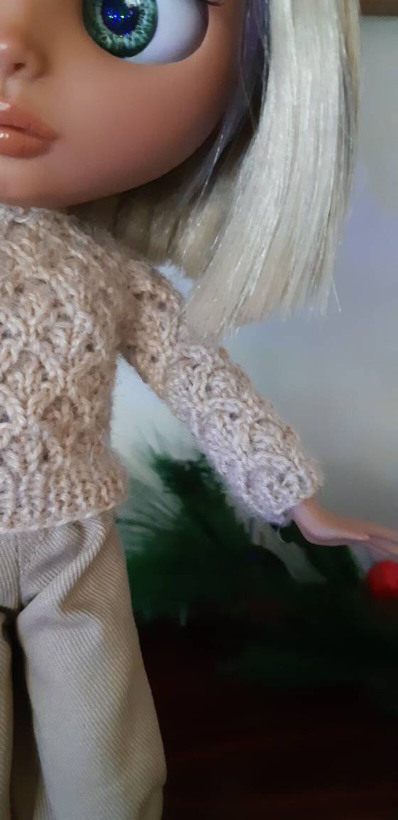 May include: A doll wearing a beige knitted sweater with a cable knit pattern and beige pants.