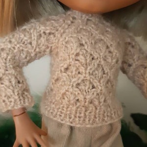 May include: A doll wearing a beige knitted sweater with a delicate openwork pattern and beige pants.