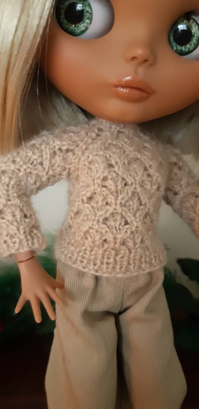 May include: A doll wearing a beige knitted sweater with a delicate openwork pattern and beige pants.