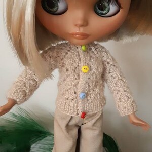 May include: A doll with blonde hair wearing a beige knitted cardigan with colorful buttons and beige pants. The doll is wearing white sneakers and has green eyes.