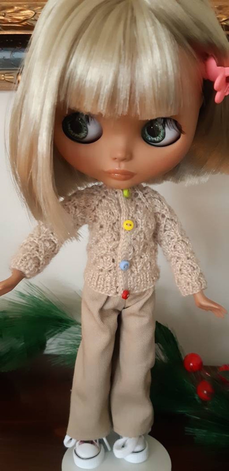 May include: A doll with blonde hair wearing a beige knitted cardigan with colorful buttons and beige pants. The doll is wearing white sneakers and has green eyes.