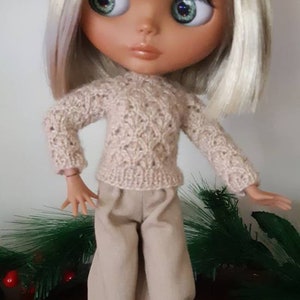 May include: A doll with blonde hair wearing a beige knitted sweater and beige pants. The doll is wearing white sneakers and is standing on a white platform.
