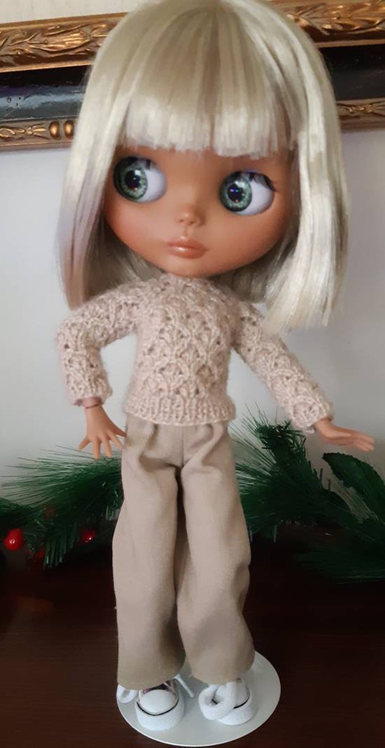 May include: A doll with blonde hair wearing a beige knitted sweater and beige pants. The doll is wearing white sneakers and is standing on a white platform.