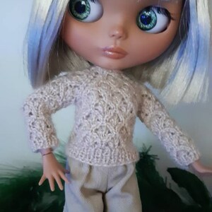 May include: A doll with white and blue hair wearing a beige knitted sweater and beige pants.