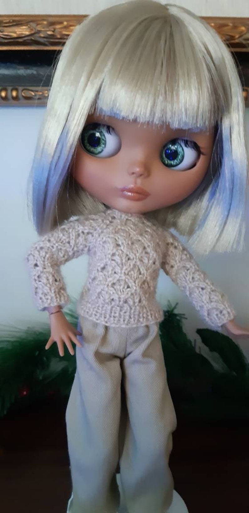 May include: A doll with white and blue hair wearing a beige knitted sweater and beige pants.