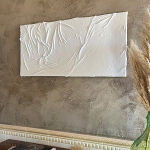 May include: White textured wall art with a raised, wrinkled surface. The art is hanging on a brown wall with a white trim.