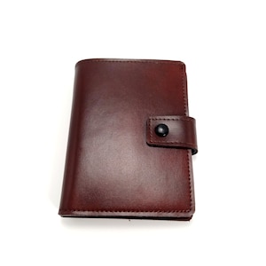 Puede incluir: Una cartera de cuero marrón con cierre de solapa y botón. La cartera tiene un borde cosido y una superficie lisa.
