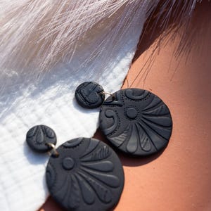 May include: A pair of black clay earrings with a floral design. The earrings are round and have a textured surface.