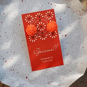 Op de afbeelding: Oranje bloemen oorknopjes op een rode kaart met het woord "Gracias!!" en "Handmade by Karla Vega" erop gedrukt. De kaart ligt op een wit en grijs gespikkeld oppervlak.