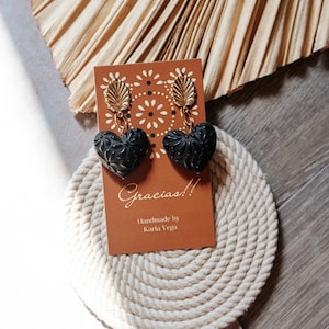 Può includere: Orecchini neri a forma di cuore con un design floreale strutturato, sospesi da accenti di foglie color oro. Gli orecchini sono esposti su una carta marrone con la scritta "Gracias!!" e il nome del produttore.