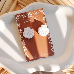 May include: White seashell stud earrings on a brown card with the brand name "HoZho" and the phrase "HECHO CON AMOR". The earrings are displayed on a white, wavy-edged dish, with a natural, textured background.
