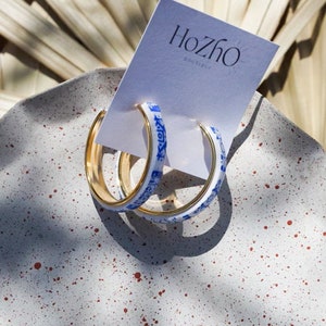 Puede incluir: Un par de pendientes de aro dorados con un diseño estampado en blanco y azul. Los pendientes están sobre una superficie moteada de blanco y rojo. El texto "HozHo Jewelry" está en una tarjeta blanca detrás de los pendientes.