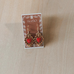 May include: A pair of gold heart-shaped earrings with red centers, displayed on a card with the text "Hozho Jewelry".