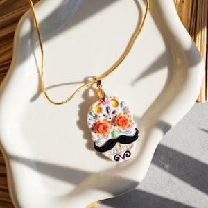 May include: A gold-colored chain necklace with a pendant shaped like a sugar skull. The pendant is white with colorful floral and decorative accents, a black mustache, and a gold-colored bail.
