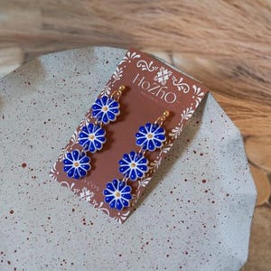 May include: A pair of blue and white floral dangle earrings with gold accents. The earrings are on a card with the text "Hozho" on it.