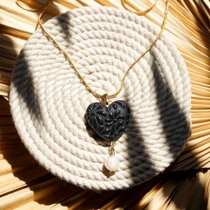 May include: A gold-toned necklace with a black heart-shaped pendant featuring a leaf design. A white pearl hangs below the pendant. The necklace rests on a white, circular woven mat, with a dried palm leaf in the background.