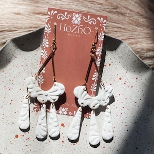 Könnte beinhalten: Weiße Keramikohrringe mit einem goldfarbenen Metallhaken und einem filigranen, komplizierten Design. Die Ohrringe haben eine geschwungene, gewellte Form mit drei tropfenförmigen Anhängern, die darunter hängen. Die Ohrringe werden auf einer weiß-rot gesprenkelten Oberfläche präsentiert.