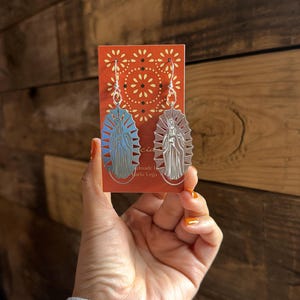 May include: Silver-toned earrings featuring a religious figure design. The earrings are held in front of a wooden background. The earrings are on a red card with the text "handmade by Karla Vega". One earring has a blue background, the other has a brown background.