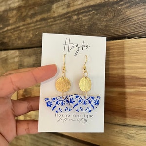 May include: Blue and white ceramic earrings with gold accents. The earrings are shaped like half circles and feature a floral design. The earrings are hanging from gold hooks.